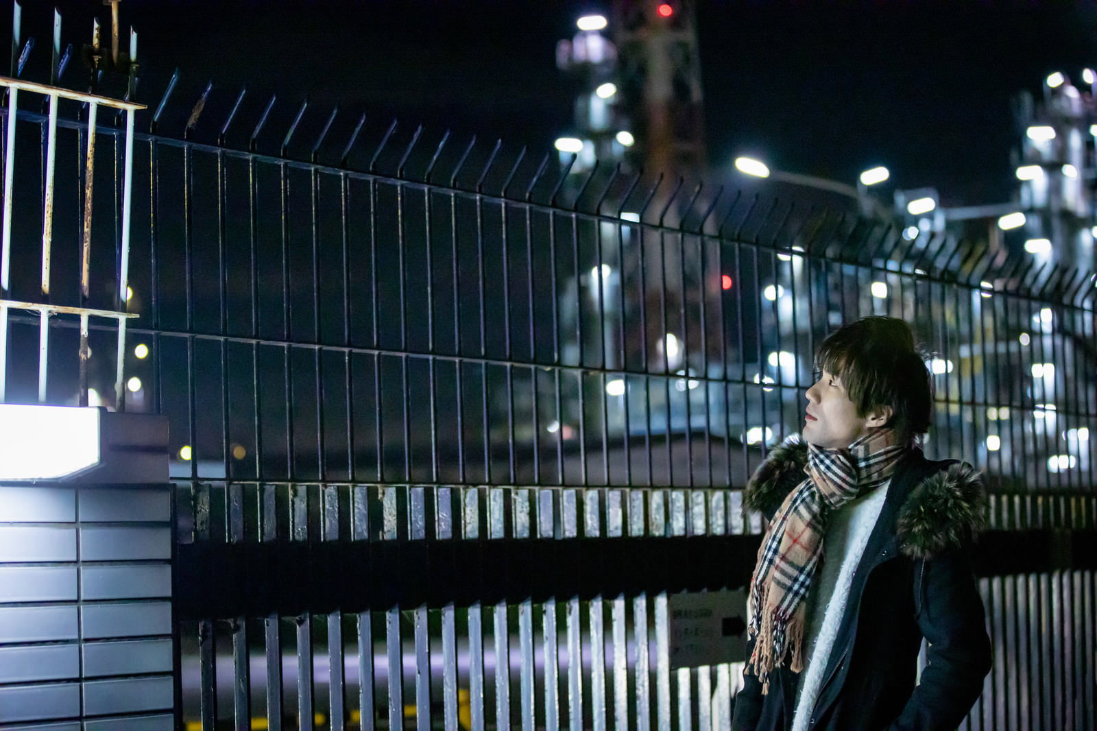 工場夜景を背景にマフラーを巻いた男性が立っている様子［モデル：村田裕章］