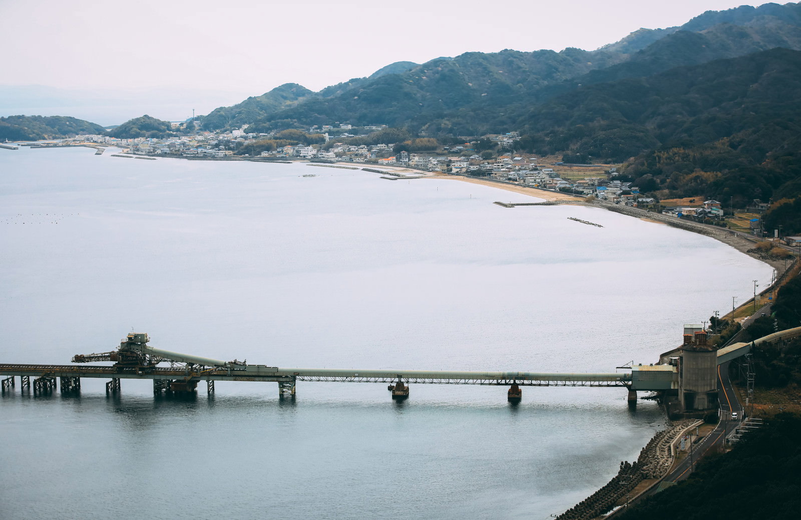 工場の建物越しに海岸沿いの漁村集落を望んだ風景