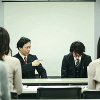 記者会見中に役員にダメ出しされる広報の写真