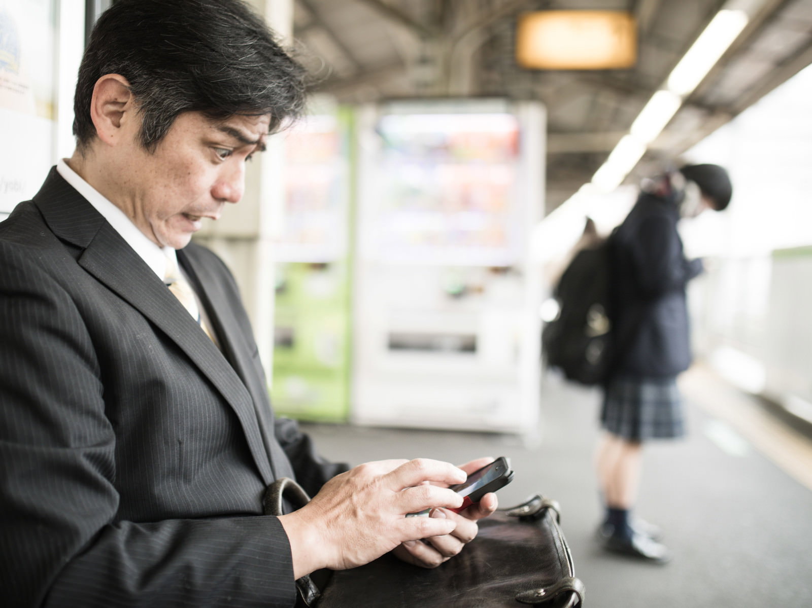 駅のホームでスマホを見て驚く会社員の男性［モデル：よたか］