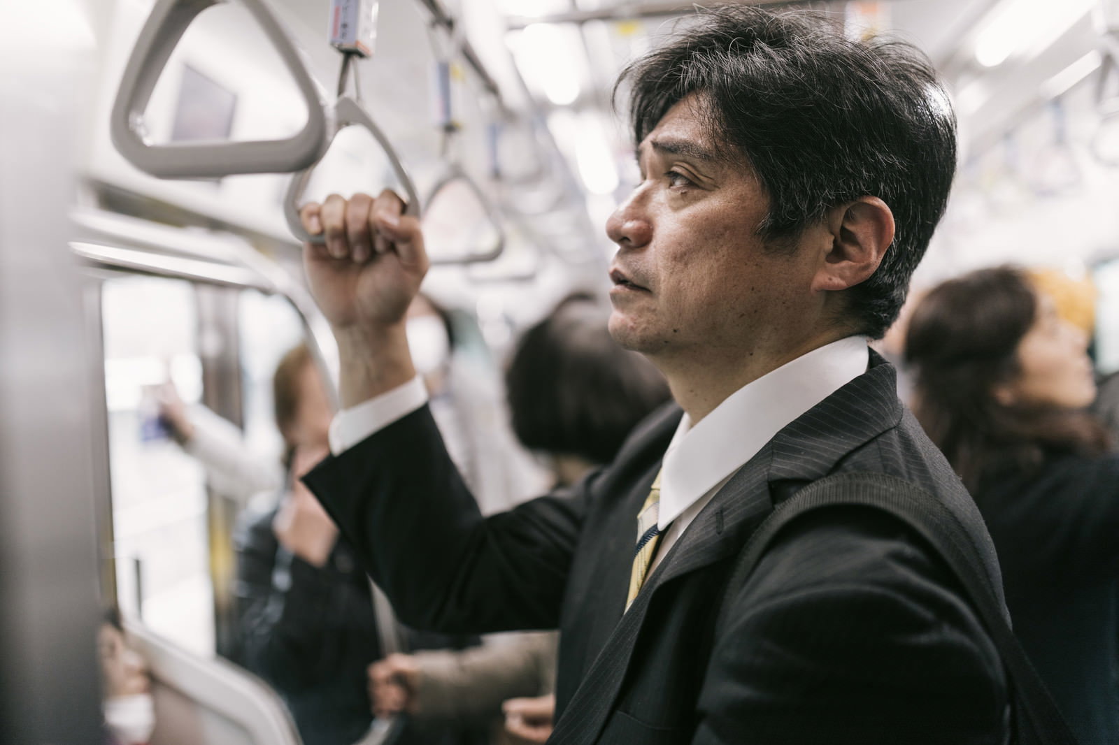 満員電車で吊革を握り立つ疲れたスーツの男性［モデル：よたか］