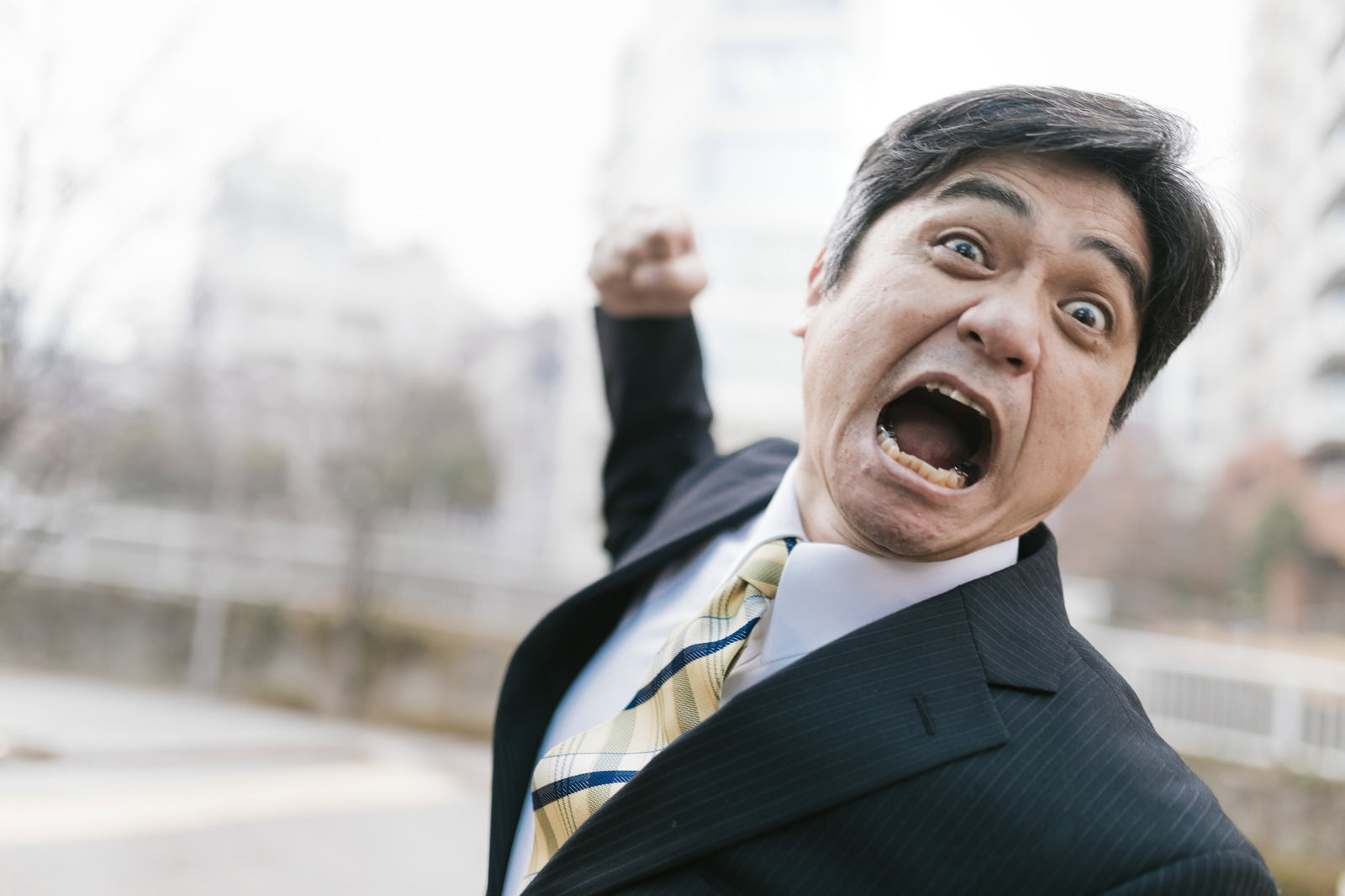 怒った表情でパンチを放つスーツ姿の男性。城市背景［モデル：よたか］