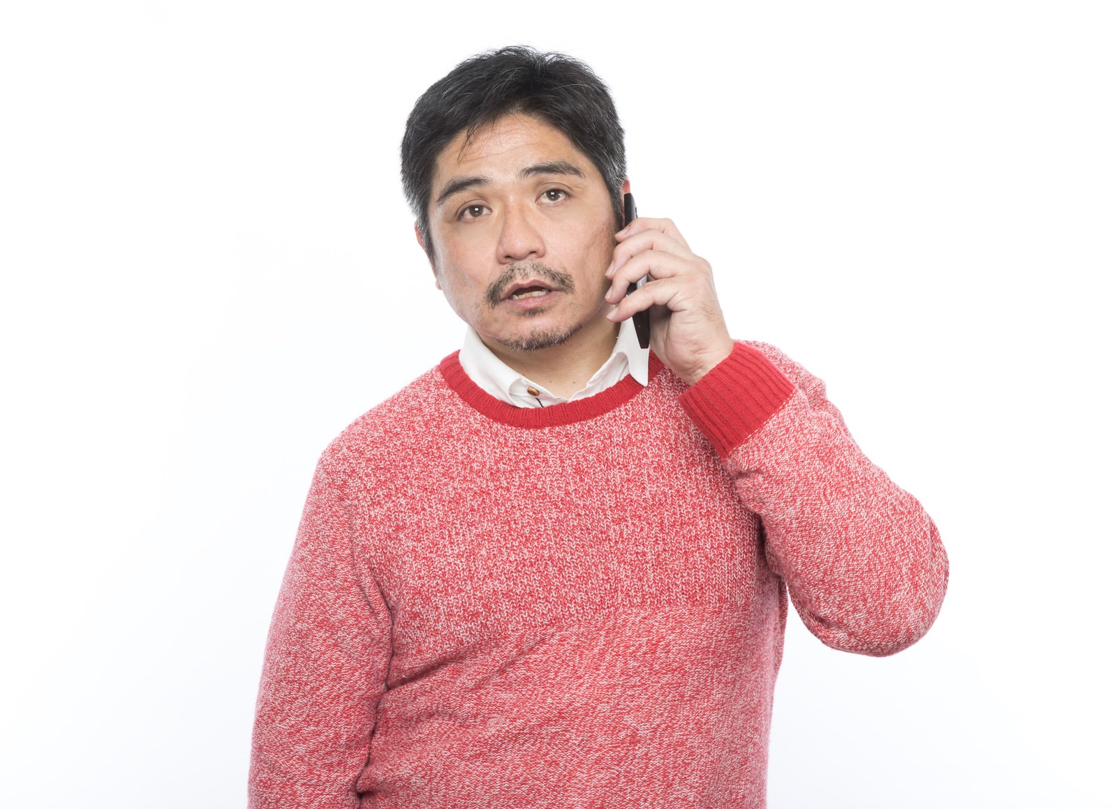 赤いセーターを着た男性がスマートフォンで電話をしながら呆れた表情をしている［モデル：よたか］