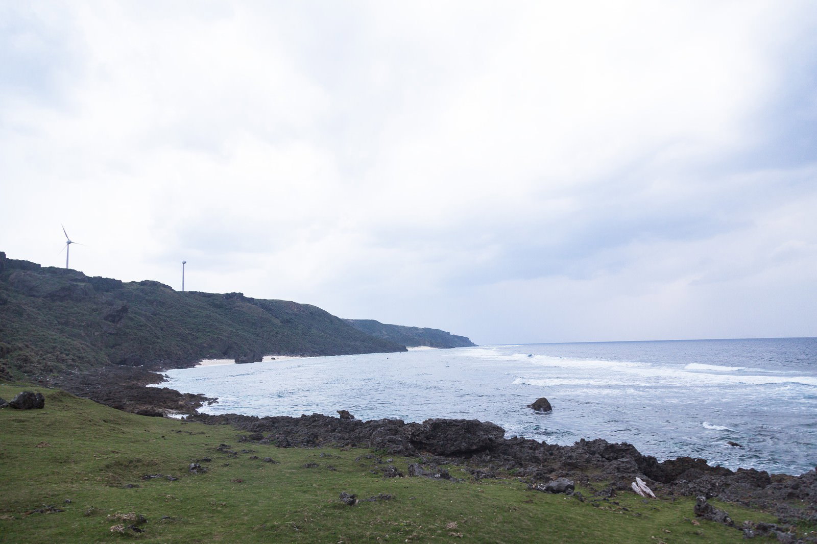 与那国島の芝生が広がる断崖の上に建つ風力発電機と、足元の黒い岩礁が波打つ青い海に囲まれた風景