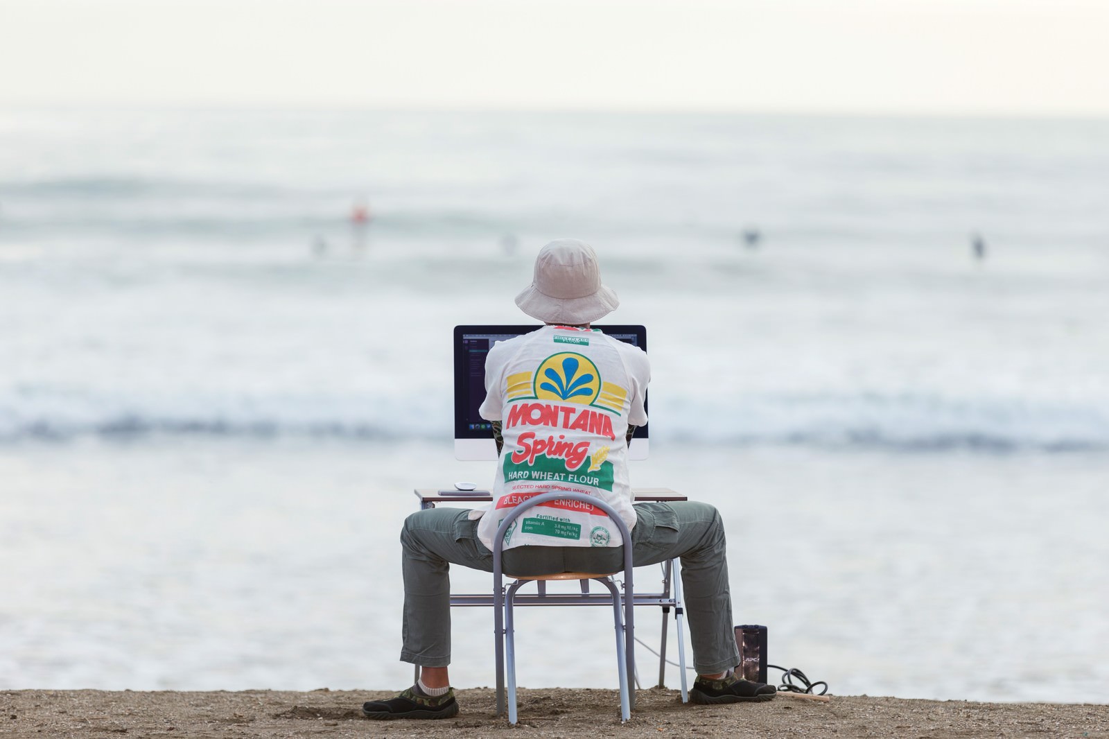 砂浜でパソコンを操作し、背後の海でサーフィンをする人々を眺めるデザイナー