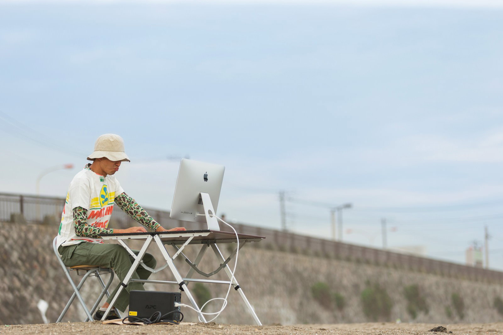 屋外で机の上のパソコンに向かって作業する男性。青空が広がる背景