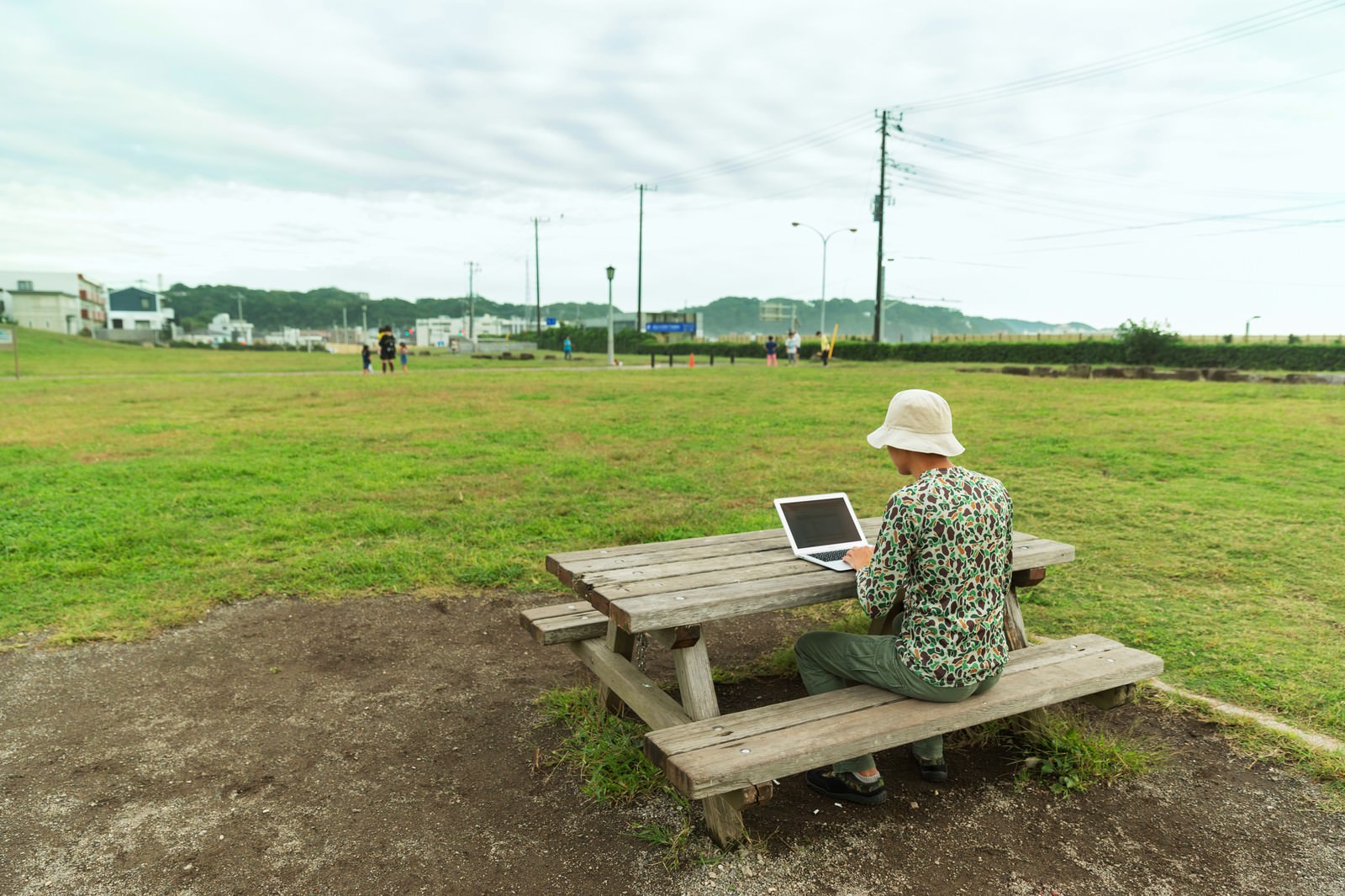 公園のベンチでノートパソコンを使って作業する白いハット姿の男性