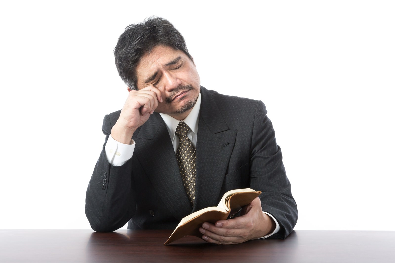 本を読みながら疲れ目で頭を抱える黒いスーツの中年男性［モデル：よたか］