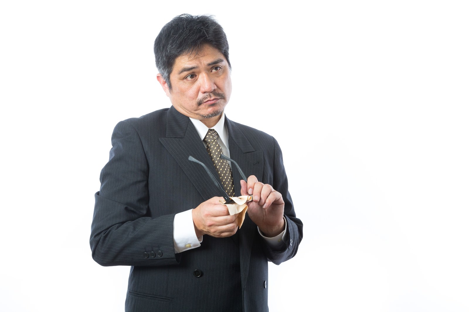 黒いスーツとドット柄ネクタイの眼鏡をかけた中年男性が、ペンを握りながら厳しい表情で指摘している［モデル：よたか］
