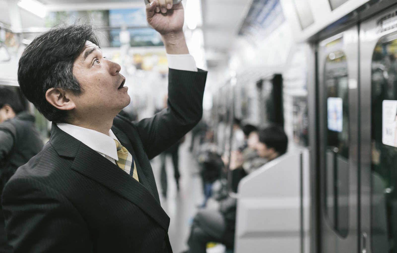 電車内で吊革を握り驚いた表情を見せるスーツ姿のサラリーマン［モデル：よたか］
