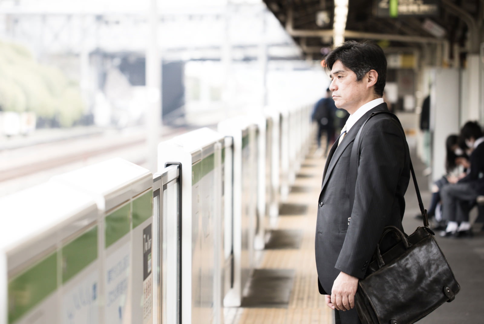 駅のホームで険しい表情を浮かべるグレースーツの中年男性会社員［モデル：よたか］