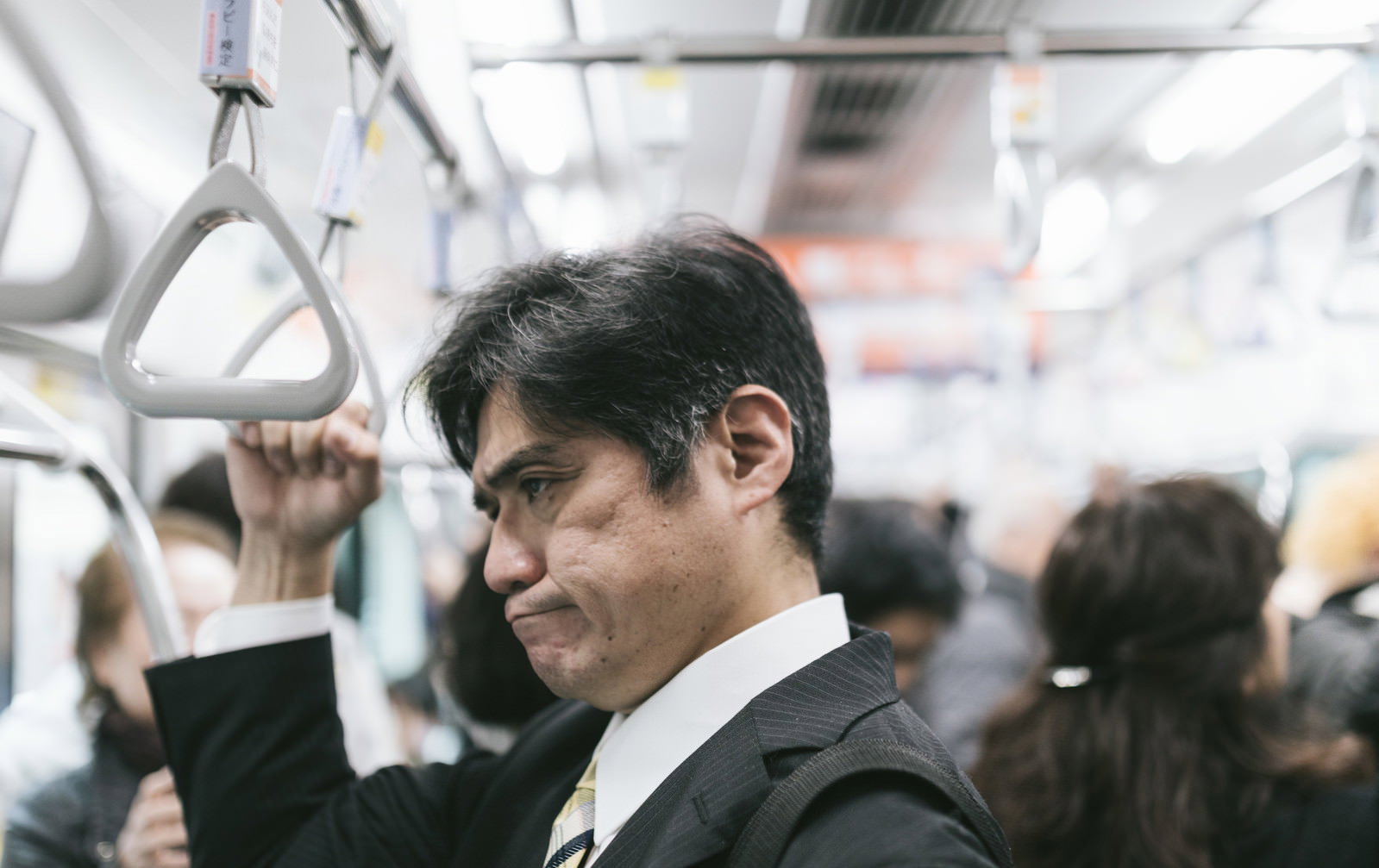 満員電車内で吊革を握り不機嫌な表情を浮かべるスーツ姿の会社員男性［モデル：よたか］