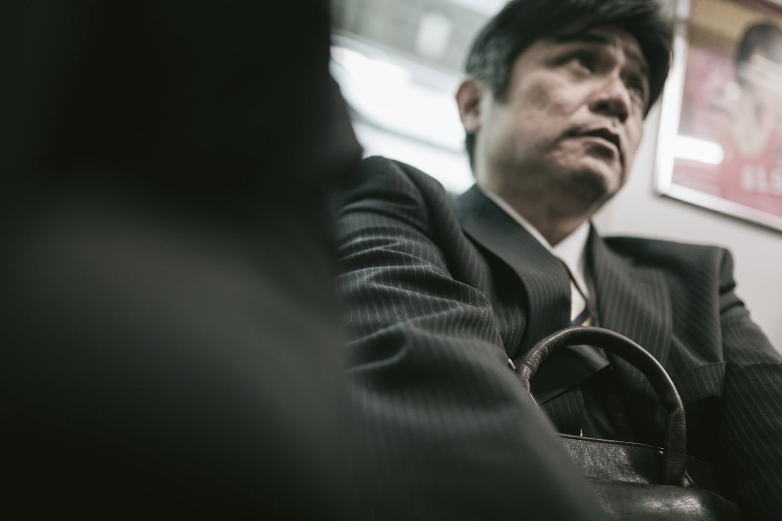 満員電車の座席に座り、周囲の乗客を気にしながら下を向く黒いスーツ姿の中年男性［モデル：よたか］