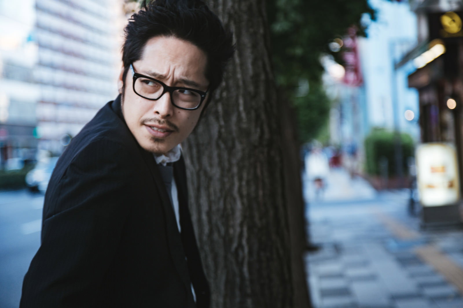 街中で後ろを振り返る眼鏡をかけた黒いスーツの男性［モデル：ゆうせい］