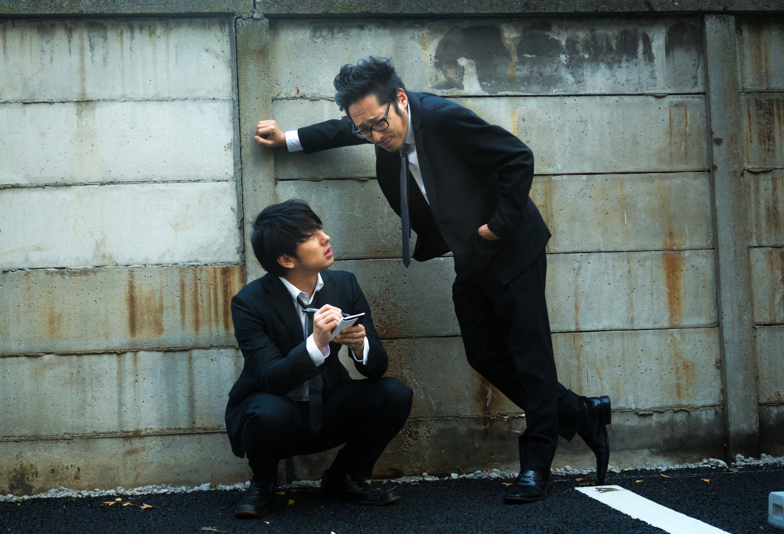 コンクリート壁前で、黒いスーツの男性が身をかがめてメモを記し、もう一人が指示する張り込み調査場面［モデル：大川竜弥 ゆうせい］