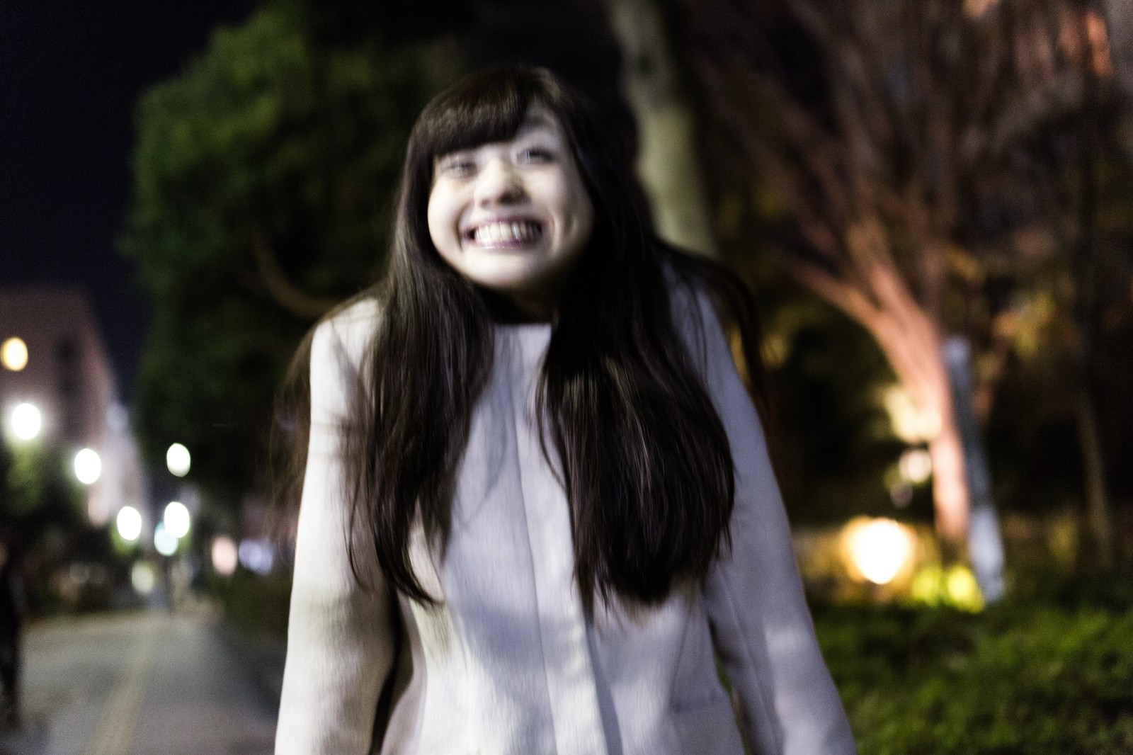 夜の公園で両手を握り満面の笑みを浮かべる紫色コート姿の女性。背景に樹木のライトアップ［モデル：河村友歌］