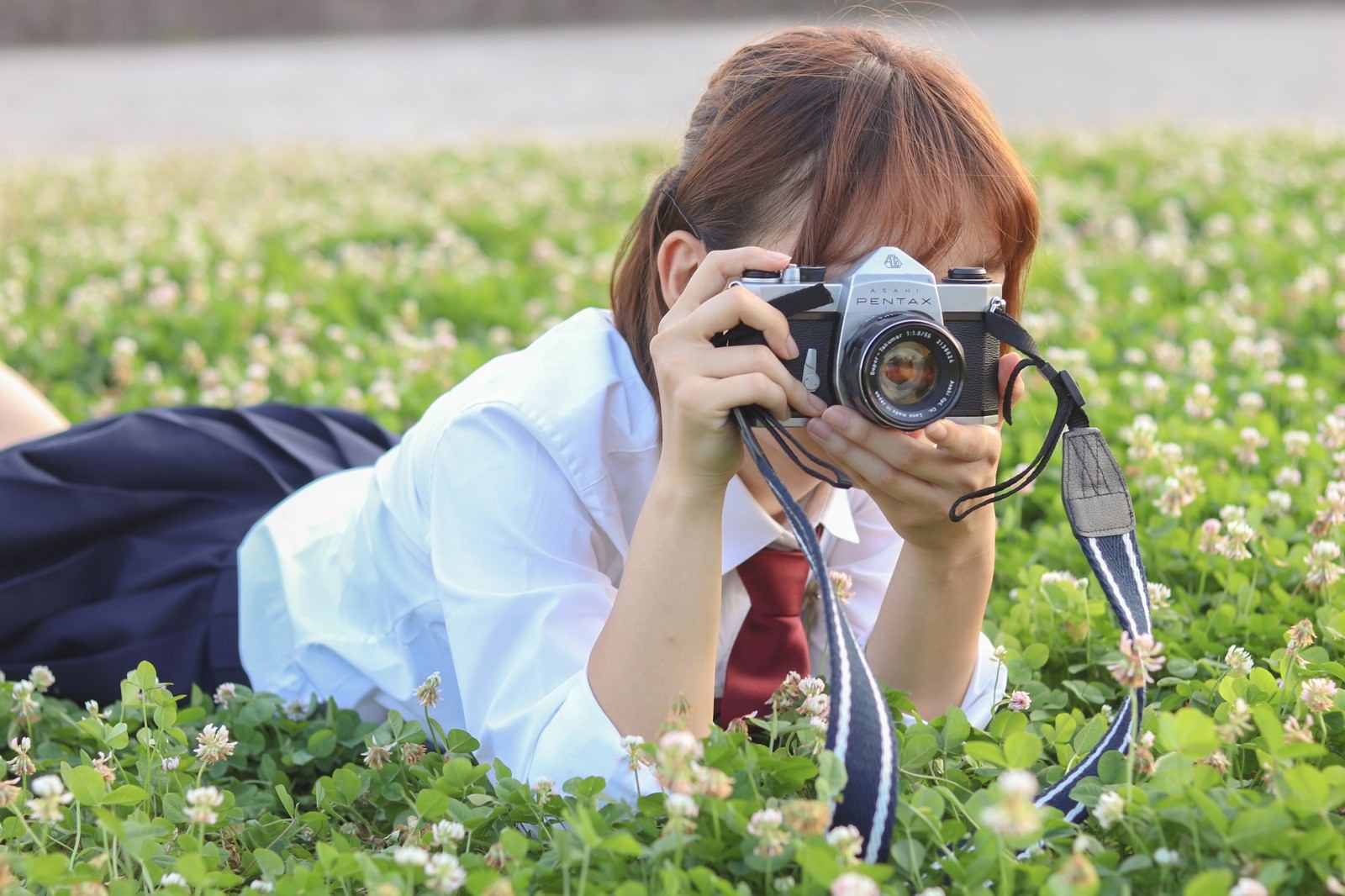 草原にうつ伏せになって一眼レフカメラを構える女子高生