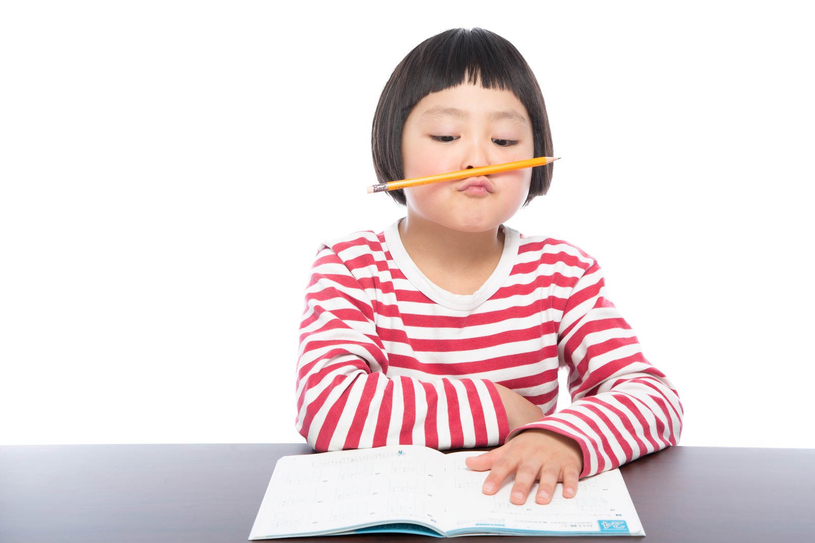 机の前で鉛筆をくわえて困った表情をする赤白ボーダーシャツの小学生［モデル：ゆうき］