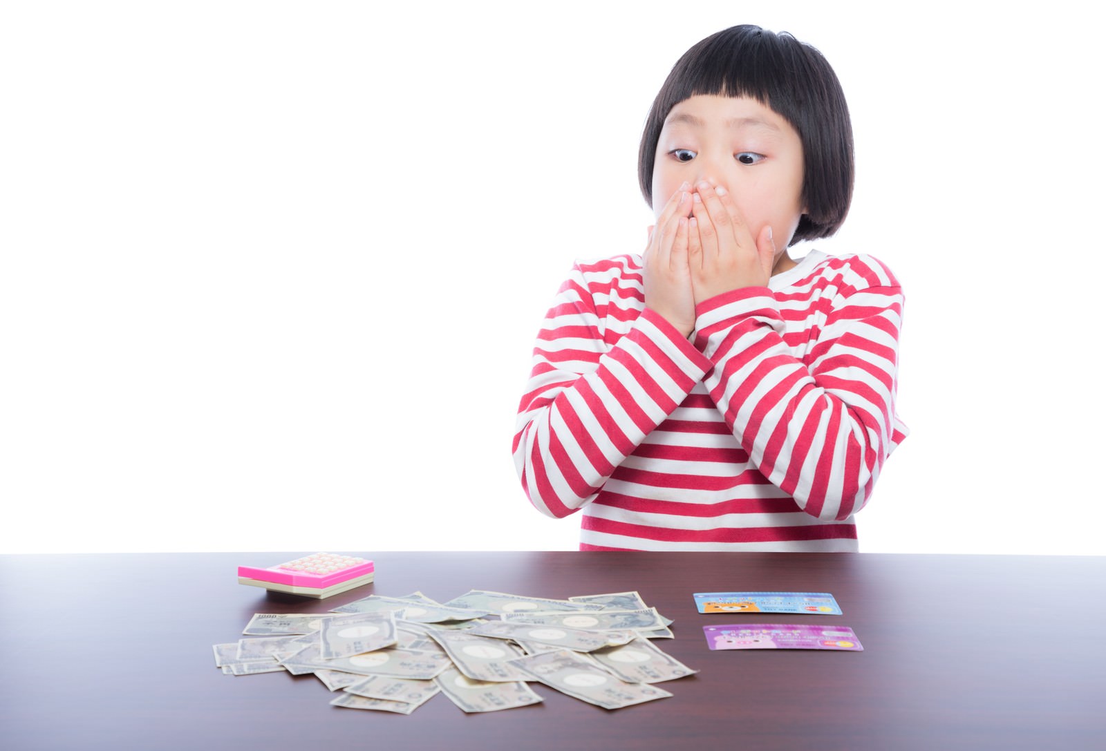 机の上の紙幣を前に、両手を口元に当てて驚く表情の前髪パッツンの女の子［モデル：ゆうき］