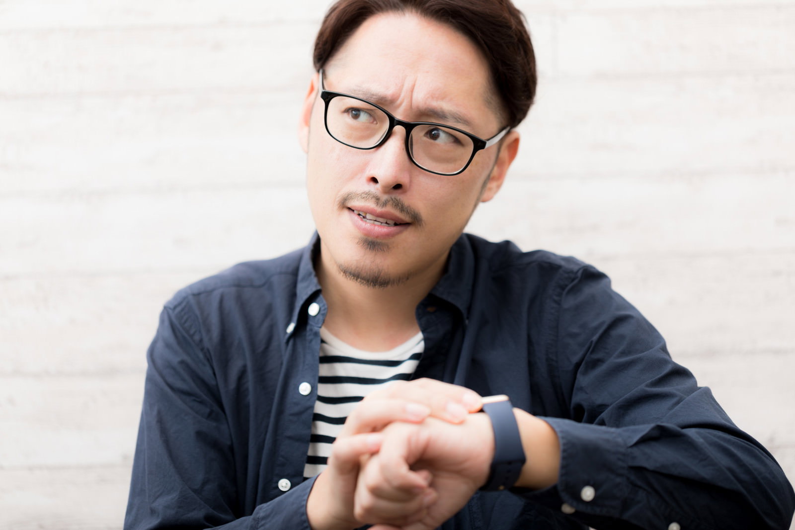 腕時計を見て時刻を確認している眼鏡をかけたデニムシャツの男性［モデル：ゆうせい］