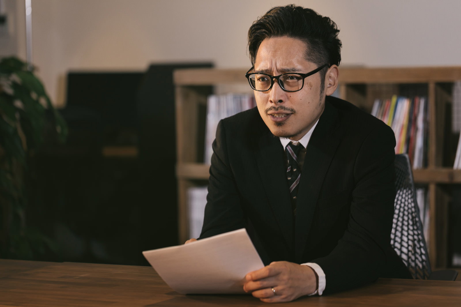 険しい表情で書類を持つスーツ姿の眼鏡をかけた男性［モデル：ゆうせい］