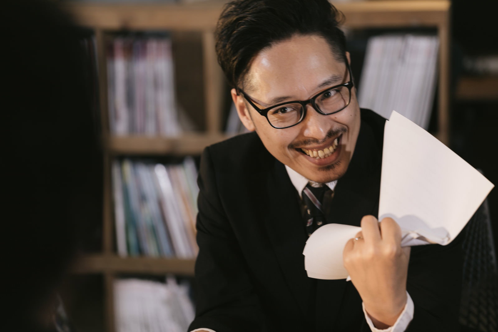 眼鏡をかけた男性がスーツ姿で書類を持ち、挑発的に笑みを浮かべている［モデル：ゆうせい］