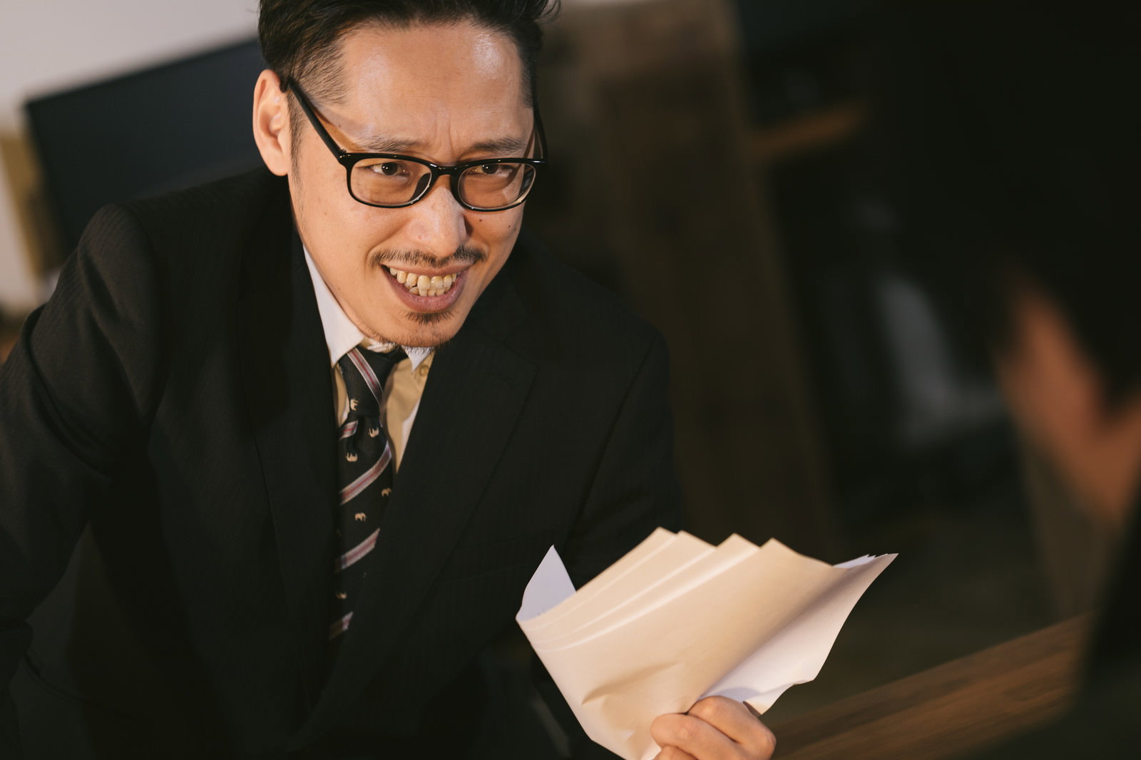 書類を持ち怪しげな笑顔で話しかけるスーツ姿の男性［モデル：ゆうせい］