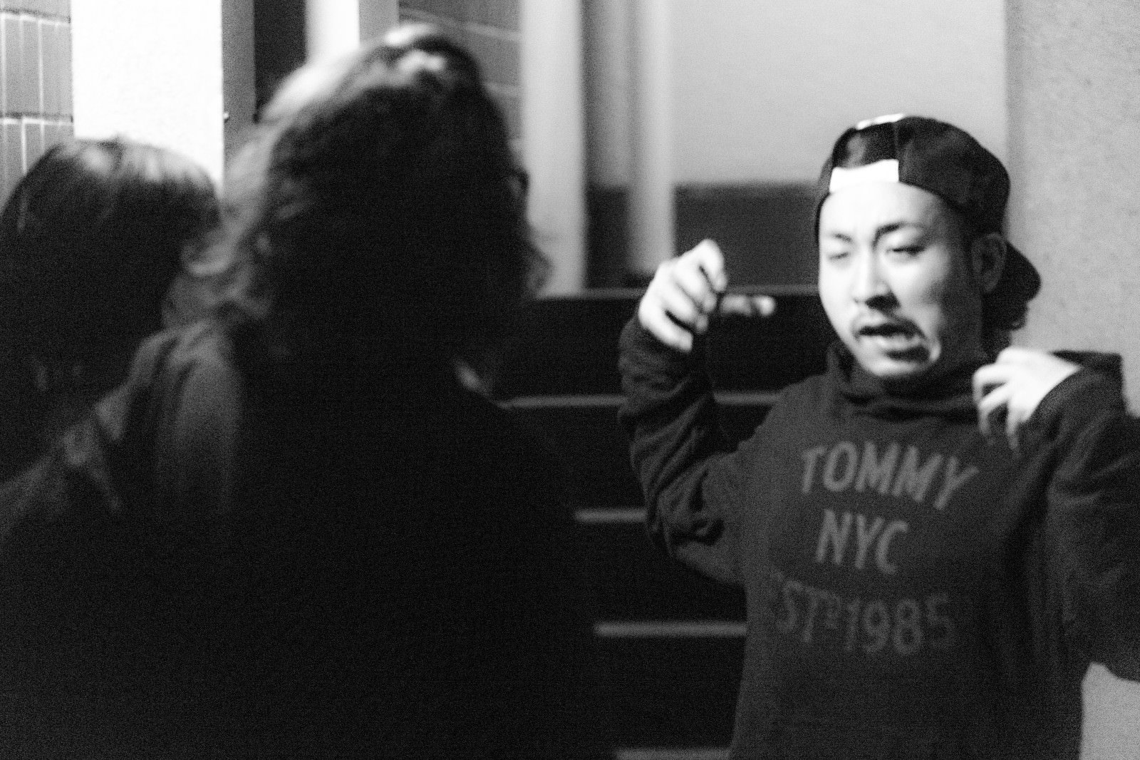 白黒室内写真。座った男性が上半身を傾け腕を上げ、口を開けている。左奥に別の人物のシルエット