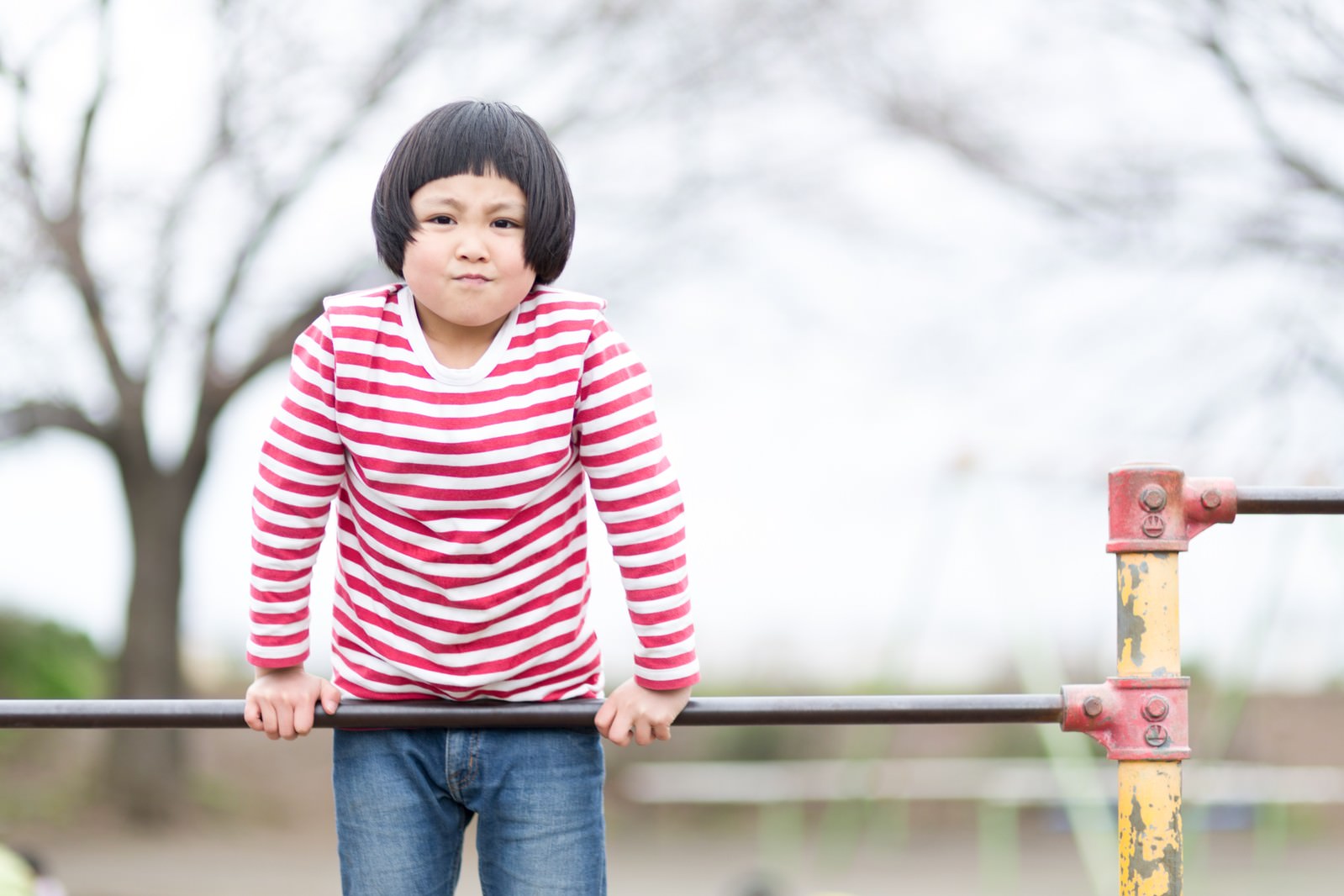 ピンクのボーダー柄の長袖シャツを着た子供が、赤い鉄棒を両手で握って練習している［モデル：ゆうき］