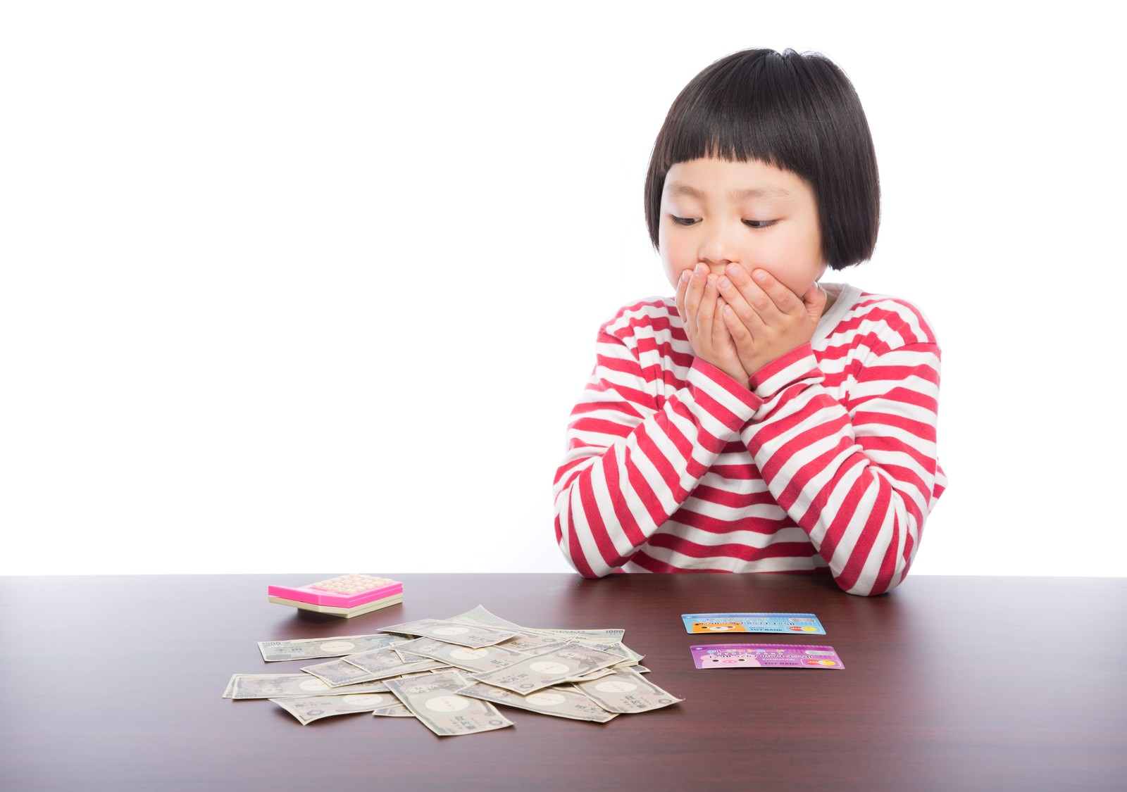 机上の現金と通帳を見つめ、両手を口に当てて驚く表情の前髪パッツン少女［モデル：ゆうき］