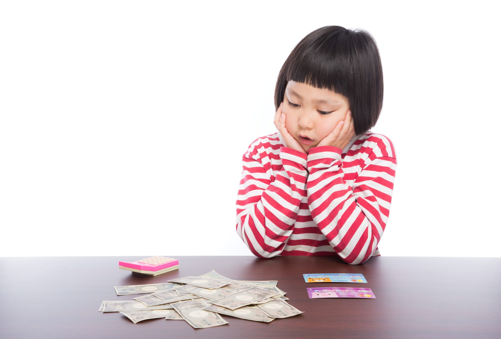 ピンク白ボーダーシャツの女の子がテーブルに座り、紙幣と通帳を前に両手で頬を支えている［モデル：ゆうき］