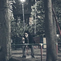 夜の公園で街灯の下のブランコに座る縞模様のシャツの眼鏡の人物の写真