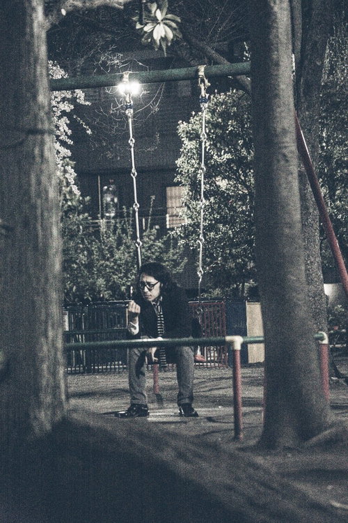 夜の公園で街灯の下のブランコに座る縞模様のシャツの眼鏡の人物