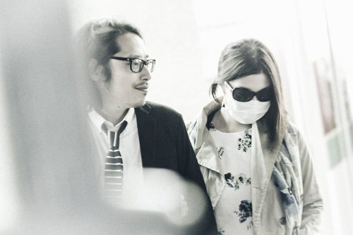 シャツの眼鏡男性とサングラスの花柄女性が並んで立つ室内写真