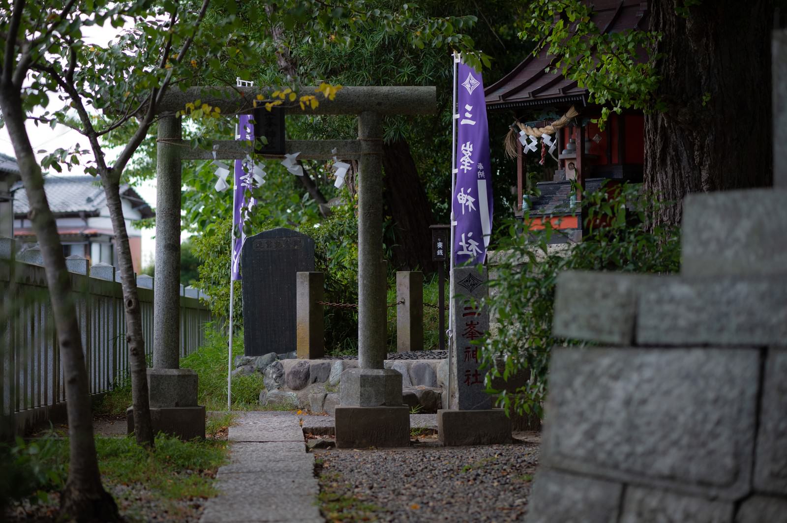 木々に囲まれた玉前神社境内の三峰神社、石の鳥居と石灯籠が見える
