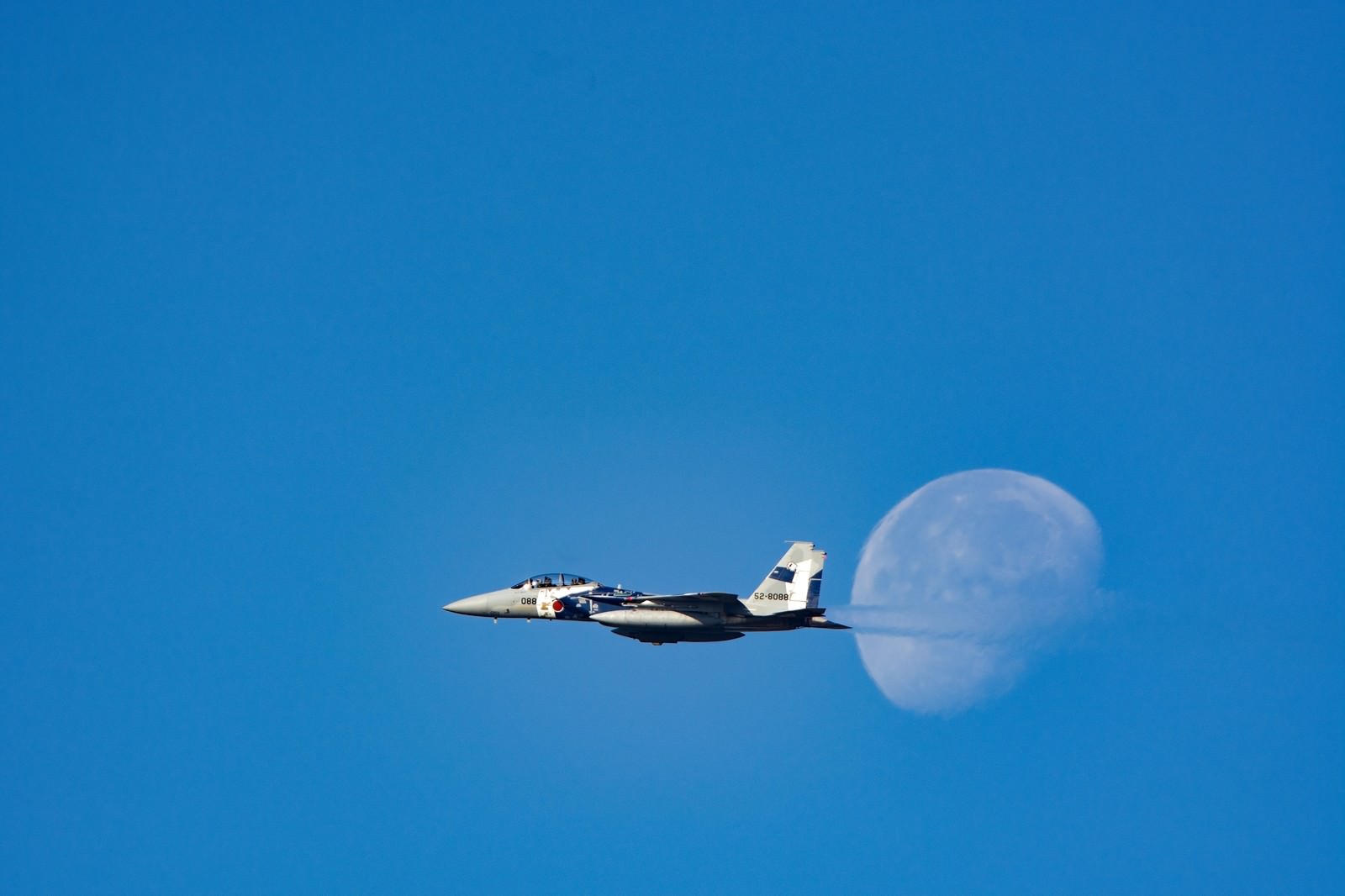 青空に浮かぶ白い月を背景に飛行するF-15戦闘機