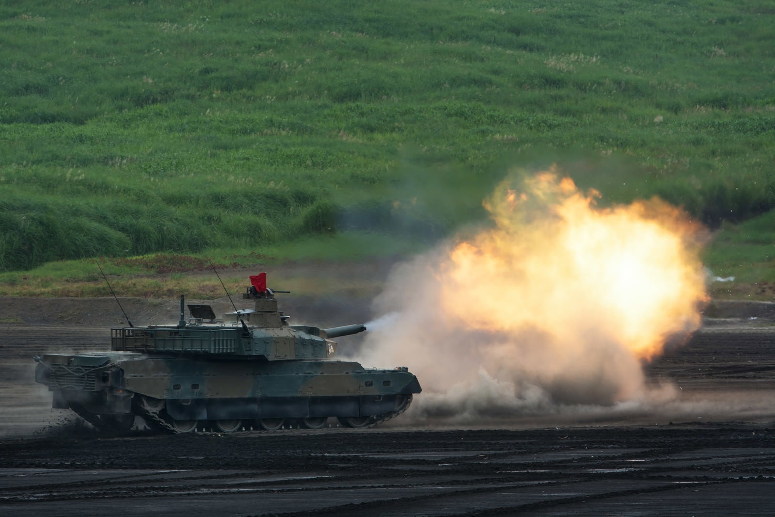 砲身から炎と白煙を噴出させながら砲撃する10式戦車。砂煙が舞い上がり背景に緑の草地が見える。