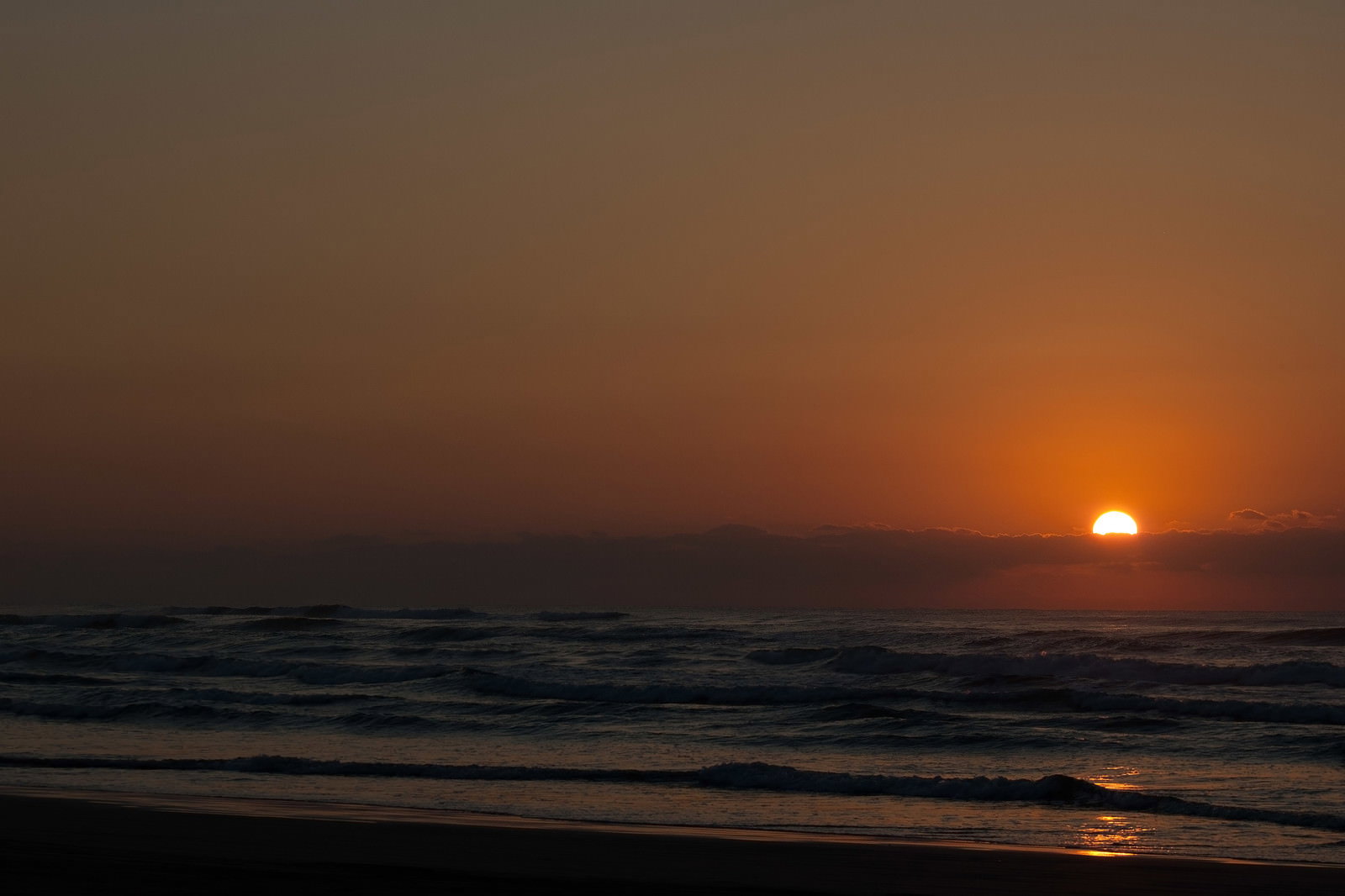 水平線に沈む夕日とオレンジ色に染まる空、波打つ海と砂浜