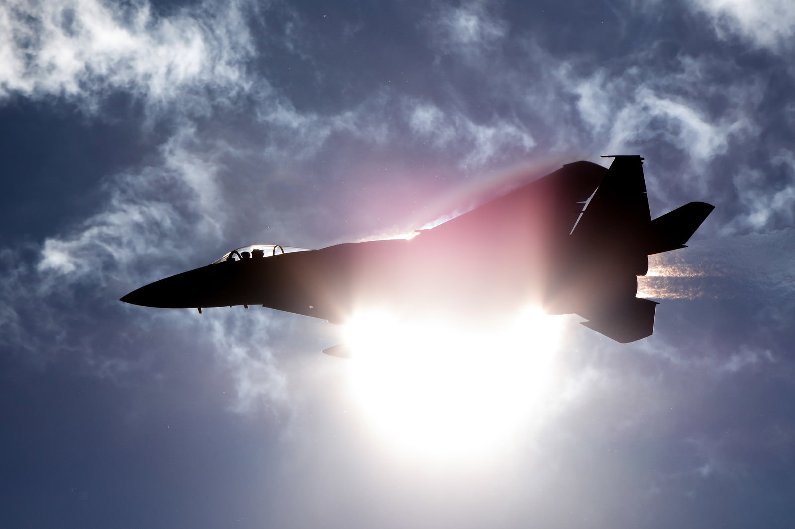 太陽を背景に逆光でシルエットになったF-15戦闘機が雲間を飛行する様子