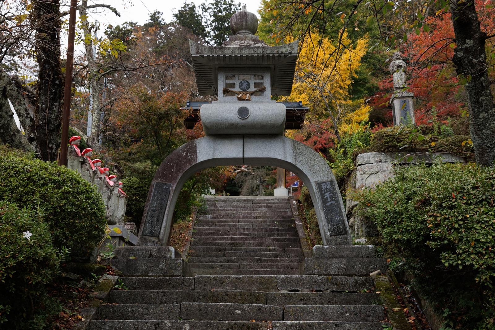 石造りの照干一隅燈籠と参道の階段、紅葉した樹木に囲まれた岩角寺の境内