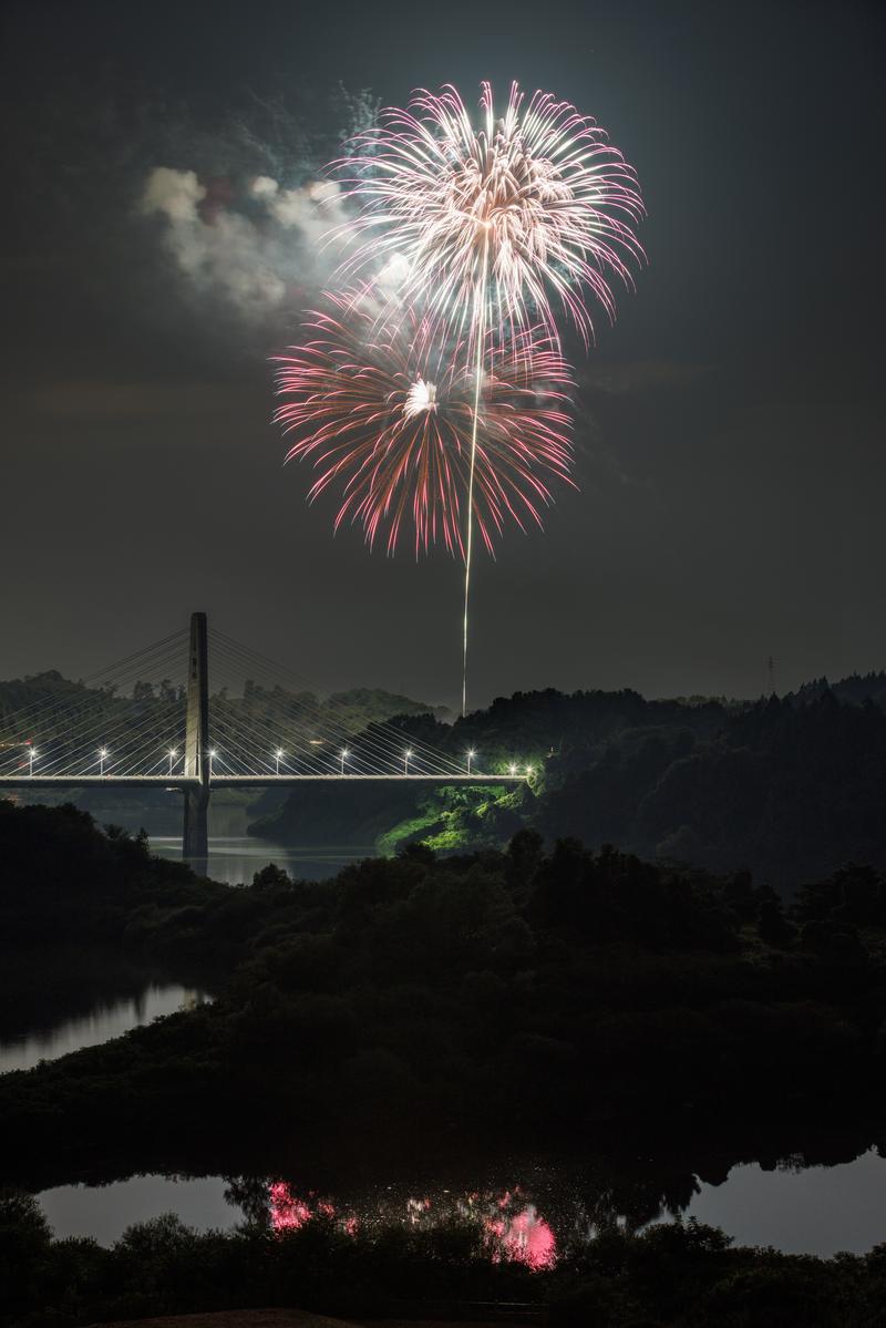 三春町の三春花火が夜空に咲き、斜張橋とその水面反射を照らす夜景
