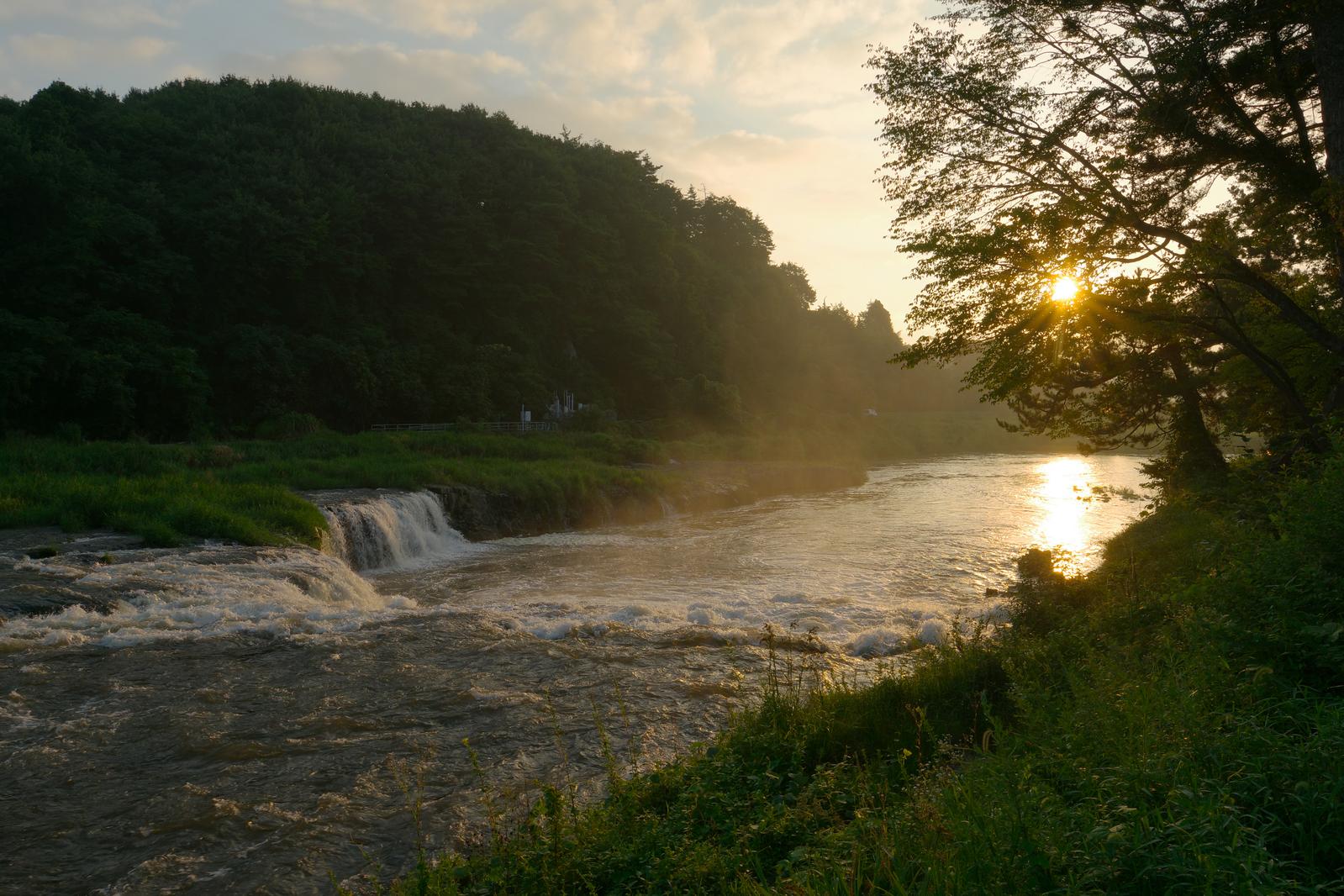 夕日を背に落ちる阿武隈川の乙字ヶ滝と緑の樹木に囲まれた渓谷