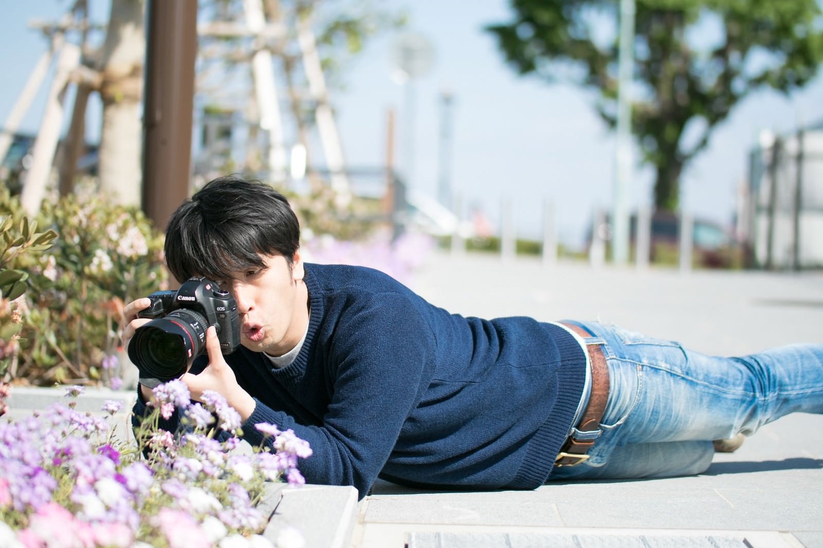 花壇の前にほふく前進のようにうつ伏せで一眼レフカメラを構えるアマチュアカメラマンの男性