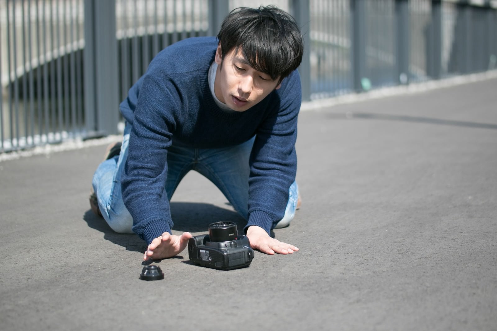 駐車場でカメラを落とし、しゃがみ込んで見つめる男性カメラマン［モデル：大川竜弥］