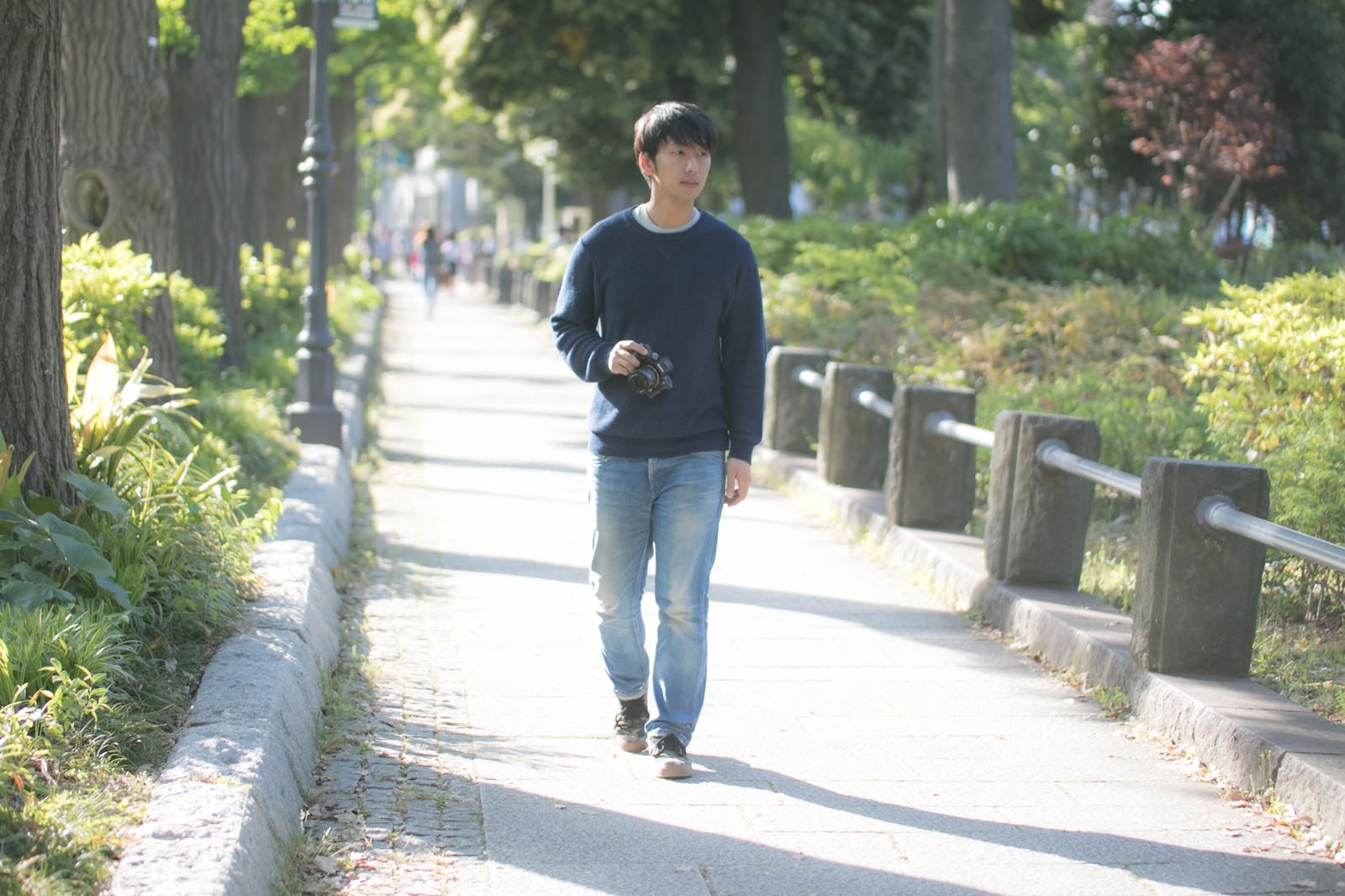 公園の並木道でカメラを持ちながら歩く男性［モデル：大川竜弥］