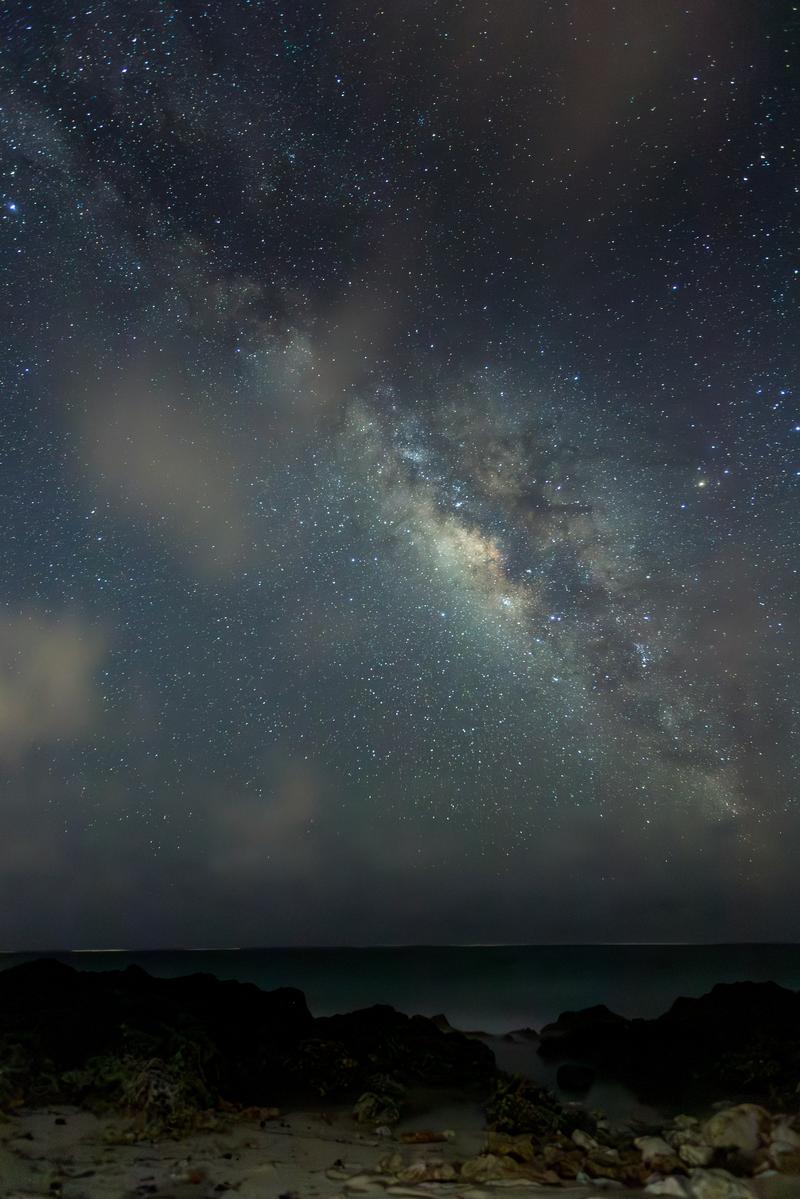 砂浜と磯の背景に、天の川が帯状に伸びる星々に満ちた夜空