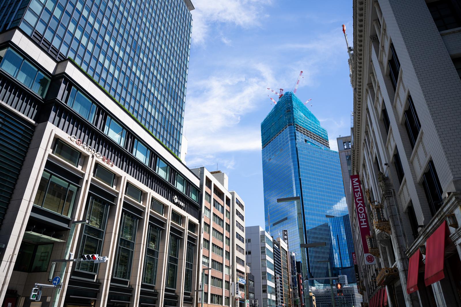 Low-angle view of Nihonbashi commercial street in Tokyo, with COREDO building on left, Mitsukoshi on right, and a blue glass skyscraper under construction at center with cranes visible against a partly cloudy blue sky.
