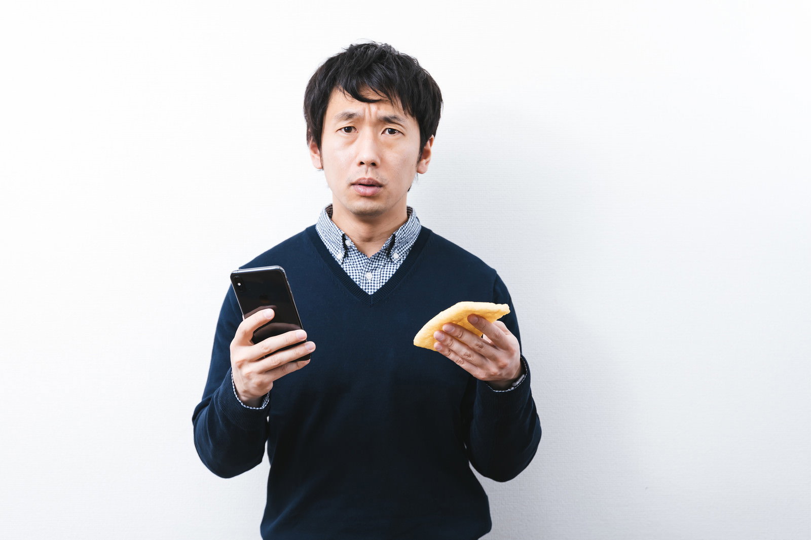 スマートフォンと油揚げを両手に持って困惑する黒いセーターの男性［モデル：大川竜弥］