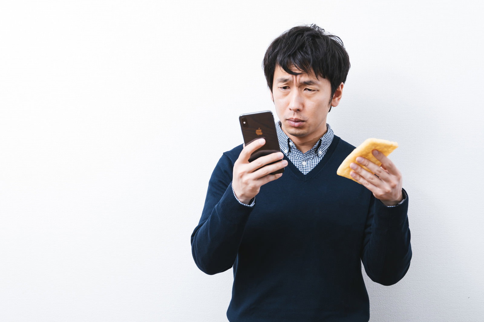 スマートフォンと油揚げを両手に持つ黒いVネックセーターの男性［モデル：大川竜弥］