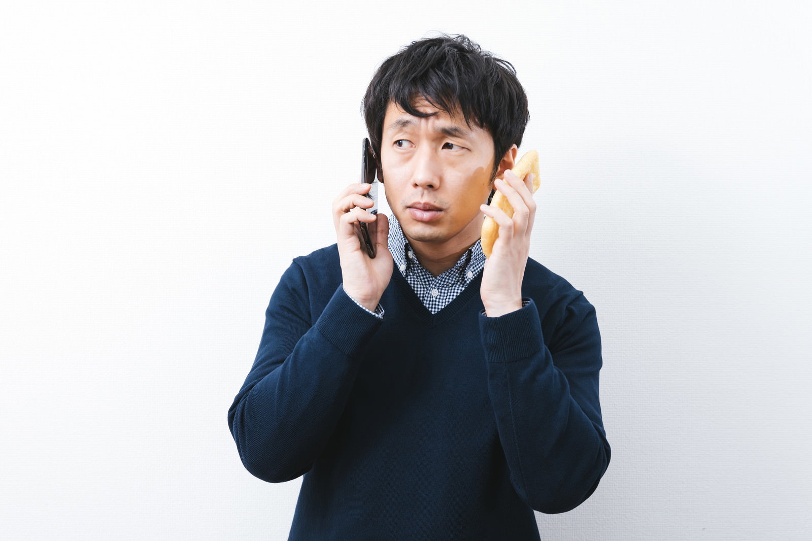 スマートフォンと油揚げを両手に持ち困惑する紺色ニットの男性［モデル：大川竜弥］